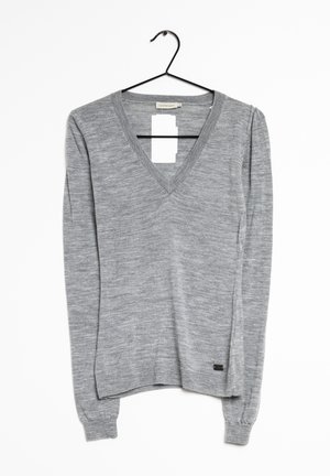Pull gris à manches longues avec col en V suspendu sur un cintre noir devant un fond blanc, avec une petite étiquette rectangulaire de marque près de l'ourlet.