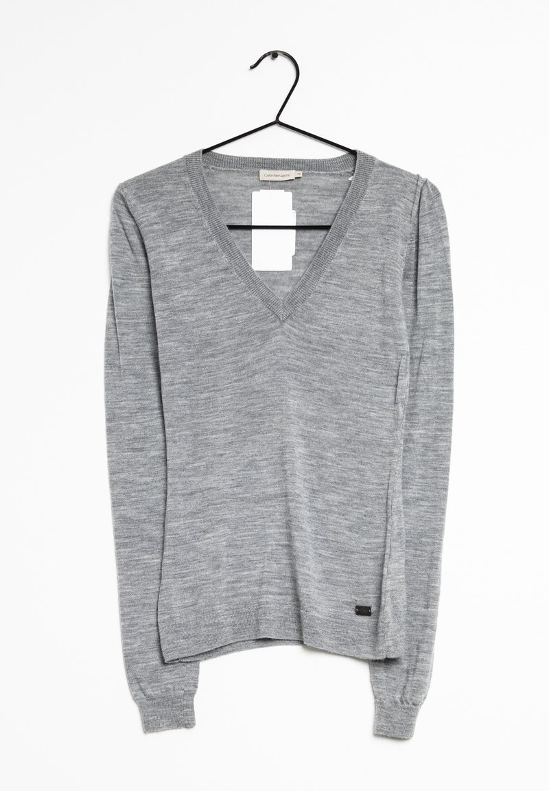 Pull gris à manches longues avec col en V suspendu sur un cintre noir devant un fond blanc, avec une petite étiquette rectangulaire de marque près de l'ourlet.
