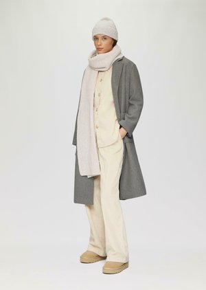 Grau-wollener Mantel über einem hellbeigen Hemd und einer hellbeigen Hose, kombiniert mit einem strukturierten, hellrosa Schal und einer passenden Mütze. Beige Schuhe runden den Look ab.