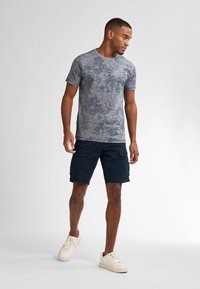 Šedé vzorované tričko s krátkým rukávem v kombinaci s námořnickými cargo šortkami. Model má na sobě krémové tenisky, což vytváří neformální, uvolněný vzhled.