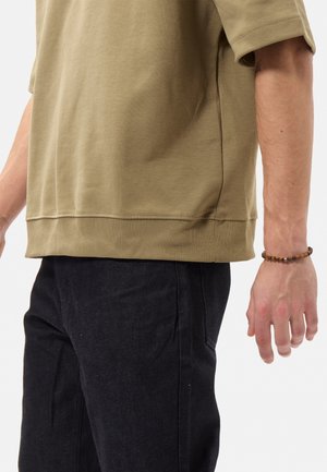 Camicia di cotone verde oliva a maniche corte con orlo a coste e spacchi laterali, abbinata a pantaloni in denim scuro e un braccialetto di perline.