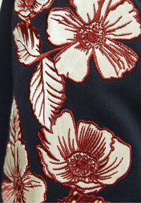 Marynarski materiał z dużymi, haftowanymi wzorami kwiatowymi w kolorze kremowym i ciemnoczerwonym, ukazujący skomplikowane ściegi i detale liści.