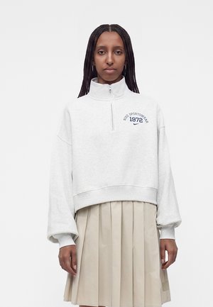 Жена с дълга плитка, носеща светлосив суичър с половин цип с "Nike Sportswear 1972" и бежова плисирана пола.