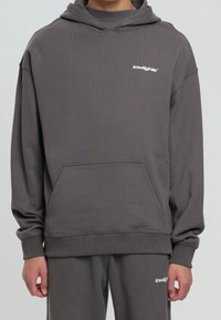 Sudadera gris con capucha, bolsillo delantero y puños acanalados. Presenta un pequeño logo blanco en el pecho. Fabricada con un material suave y texturizado.