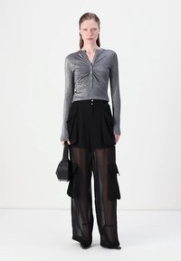 Metallic grey long-sleeve top with central ruching and button detail, paired with sheer black wide-leg trousers featuring front pockets.