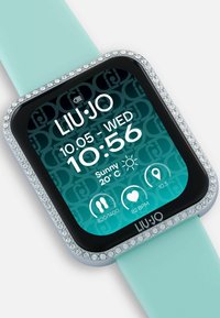 Štvorcové smart hodinky s bledotyrkysovým silikónovým pásikom, ozdobené jasnými kryštálmi okolo displeja. Digitálne rozhranie zobrazuje čas a počasie.