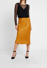 Černá blůza s dlouhým rukávem a puntíky, vrstvená přes hořčicovou saténovou midi sukni, doplněná černými vysokými podpatky.