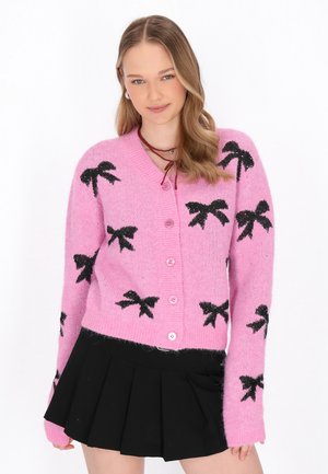Różowy sweter z dzianiny z wzorem czarnych kokardek, okrągłym dekoltem i zapięciem na guziki. Miękka tekstura z lekko puszystym wyglądem.