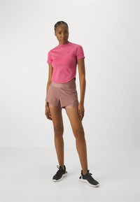Haut rose à manches courtes avec un design texturé et un logo, associé à un short rose clair avec une taille ajustée et un style superposé. Chaussures de sport noires.