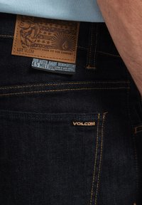Jean en denim bleu foncé avec une patch en cuir affichant un design graphique, des coutures contrastées et un petit logo "VOLCOM" sur la poche.