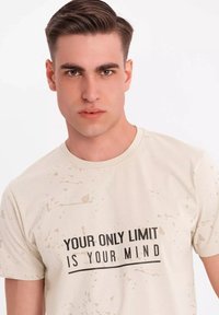 Beige t-shirt med sprøjte mønstre; har et iøjnefaldende sort tekstgrafik med teksten "DIN ENESTE BEGRÆNSNING ER DIN HJERNE" på forsiden.