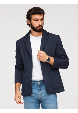 Man met donker krullend haar en baard, die een marineblauwe blazer, wit T-shirt, blauwe spijkerbroek en een zilveren polshorloge draagt en zijn jas rechtzet.