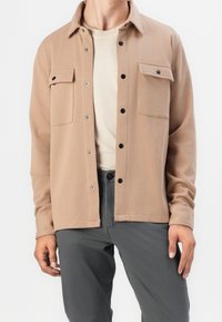 Brun knäppjacka med två bröstfickor, silverfärgade tryckknappar och en jämn textur, bärs över en ljusbeige t-shirt och grå byxor.