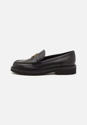 Mocassins en cuir noir avec une semelle épaisse et basse. Présentent un design piqué et un accent en métal doré sur le dessus. Texture lisse.
