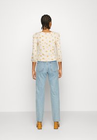 Top branco com padrão floral amarelo, mangas bufantes; combinado com jeans azul claro e sapatos de salto em tom carne. Fundo branco liso.