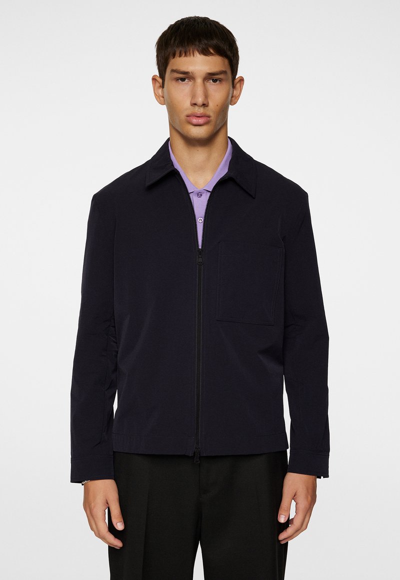 Giacca navy con zip frontale, colletto, taschino sul petto e maniche lunghe. Indossata sopra una polo viola chiaro.