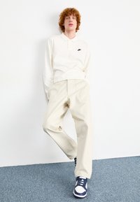 Polo de cor bege claro com colarinho e logotipo preto, combinado com calças de cor creme e ténis azul e branco, em pé sobre um tapete azul.