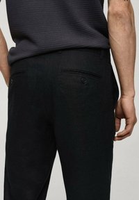 Pantaloni neri con una finitura texturizzata, dotati di due tasche posteriori con bottoni e un design aderente. Il tessuto appare leggero e informale.
