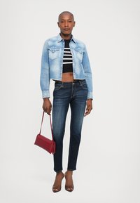 Woman wearing cropped denim jacket, striped black and white crop top, dark skinny jeans, brown heels, holding red handbag by side.