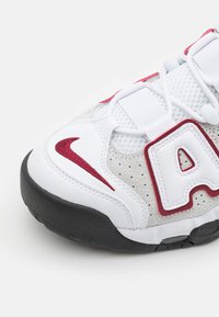 Vitt idrottssko med ovandel i mesh, röda detaljer, djärvt "A"-logotyp och en texturerad grå gummisula med en framträdande swoosh-detalj.