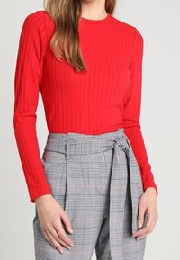 Donna che indossa un top a maniche lunghe a coste rosso e pantaloni grigi a quadri a vita alta con una cintura di tessuto legata in vita.