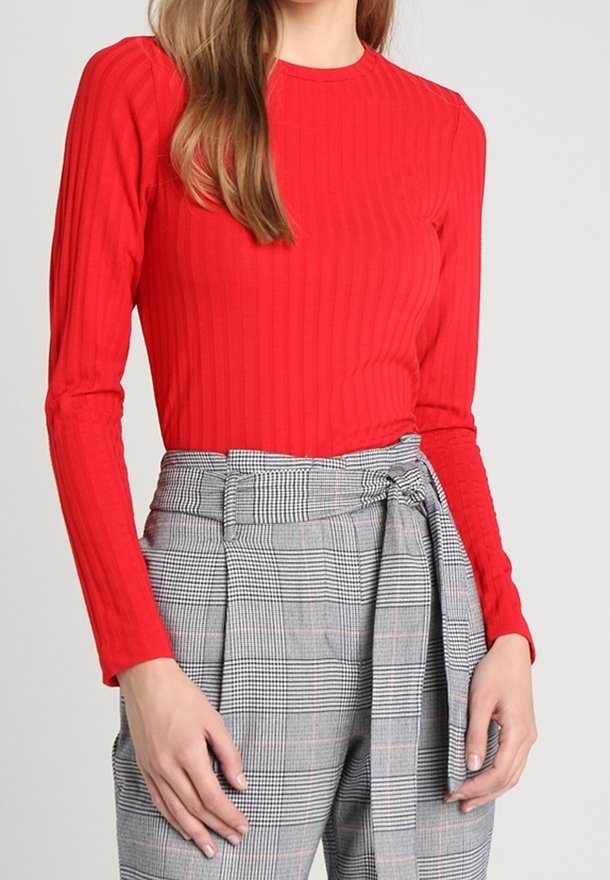 Donna che indossa un top a maniche lunghe a coste rosso e pantaloni grigi a quadri a vita alta con una cintura di tessuto legata in vita.