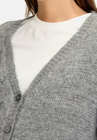 Szary dzianinowy kardigan z dekoltem w serek, wykonany z tkaniny o fakturze, z dwoma czarnymi guzikami. Nałożony na białą koszulę.