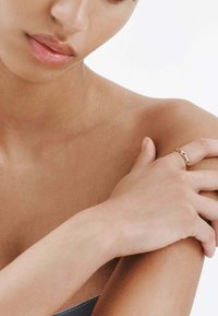 Anillo de cadena de oro en un dedo con tonos de piel suaves y claros, mostrando un primer plano de una mano descansando sobre un hombro. Fondo minimalista.