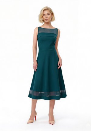 Kvinde iført ærmeløs mørkegrøn midi kjole med gennemsigtigt halsudskæring og skørt, poserer med hænderne langs siden og nøgen farvede hæle.