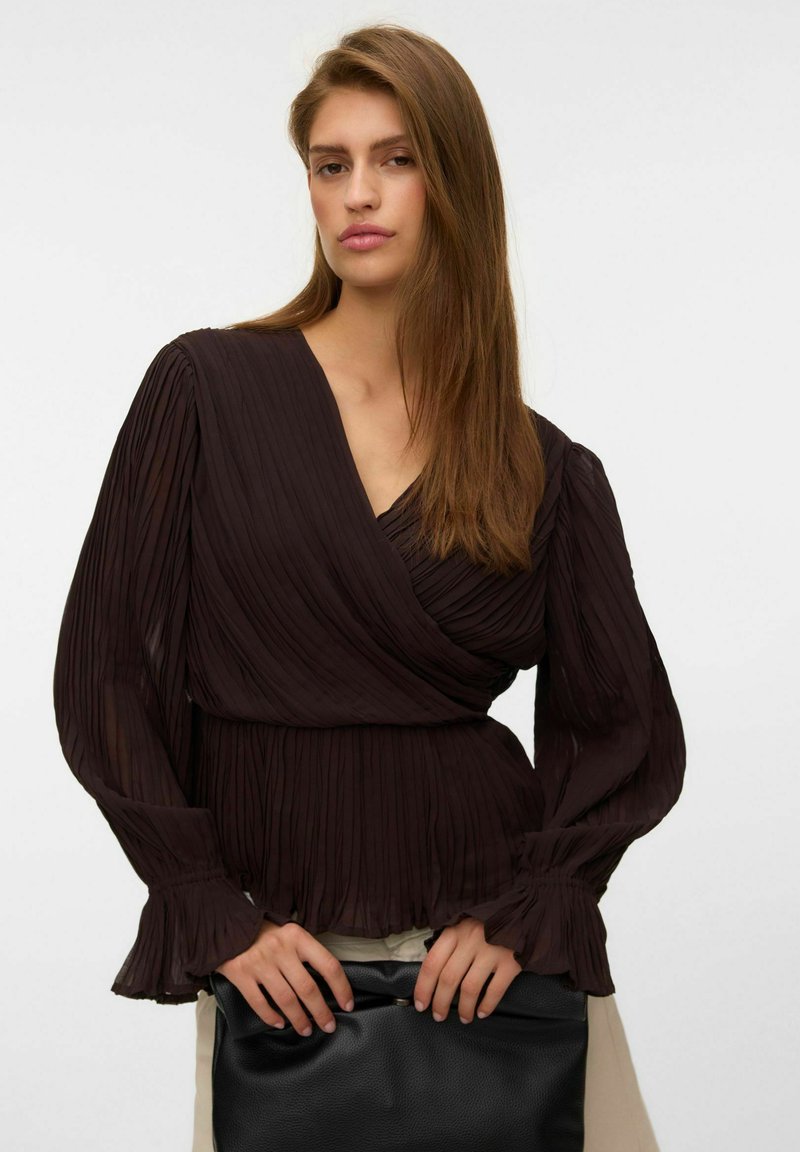 Femme aux cheveux bruns lisses porte un chemisier portefeuille plissé, à manches longues et foncé, tenant une grande pochette noire devant un fond uni.