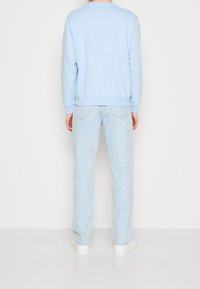 Sweat-shirt bleu clair avec poignets côtelés, associé à un jean en denim délavé clair. Baskets blanches visibles en bas. Design simple et décontracté.