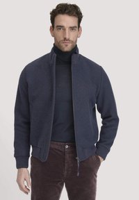 Man in een marineblauwe ritsjack over een zwarte coltrui en een paar bruine corduroybroek, staande tegen een effen lichte achtergrond.