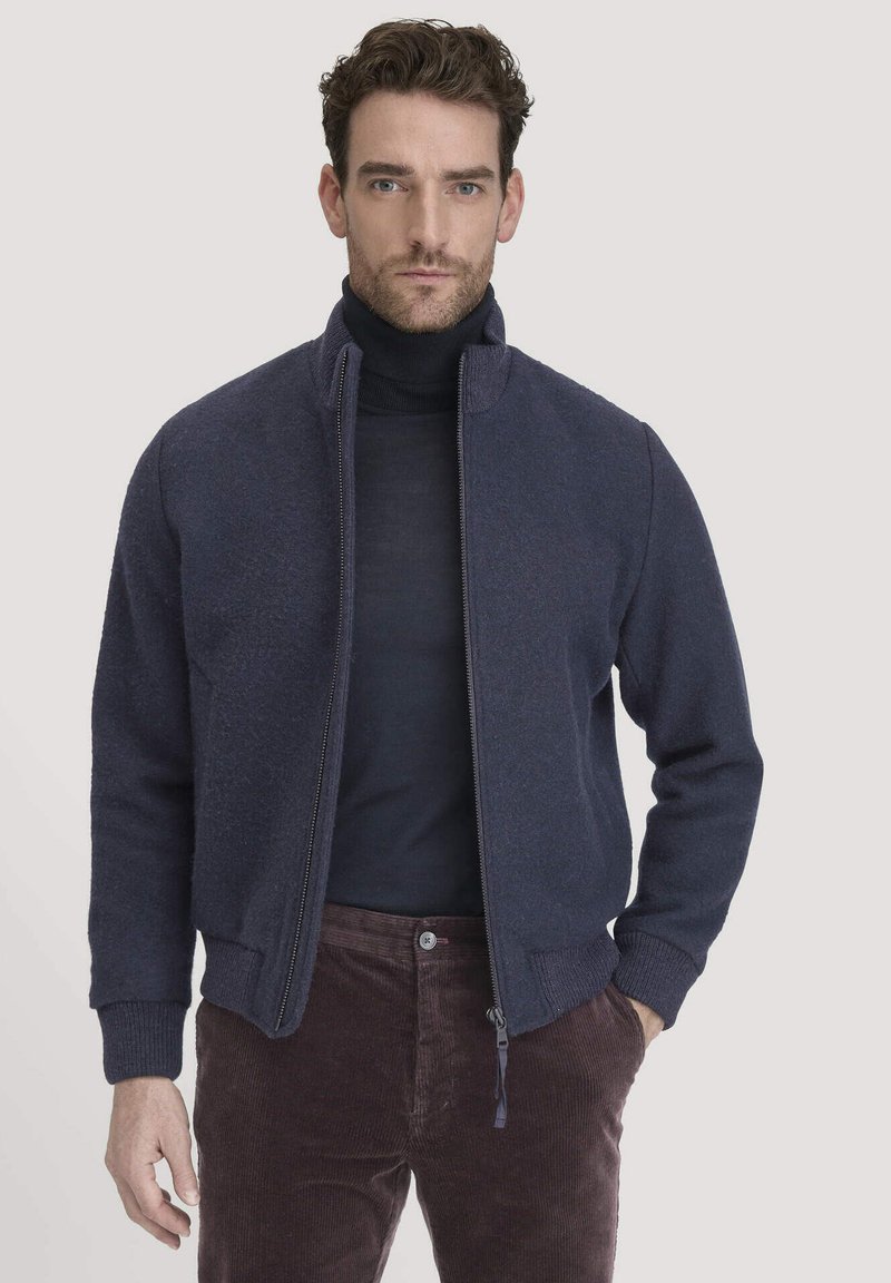 Man in een marineblauwe ritsjack over een zwarte coltrui en een paar bruine corduroybroek, staande tegen een effen lichte achtergrond.