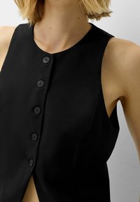 Svart ärmlös topp med hög halsringning, knappstängning fram och subtil textur. Designen är figurnära med rena linjer och inga synliga mönster.