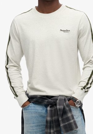 Chemise crème à manches longues avec des rayures vertes sur les manches, petit logo sur la poitrine et un tissu texturé. Portée avec un jean bleu clair.