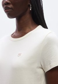 Kremowy t-shirt z krótkim rękawem, okrągłym dekoltem i haftowanym logo na piersi. Miękka tkanina, prosty fason i luźne dopasowanie.
