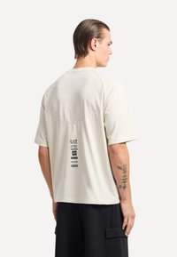 T-shirt beige chiaro con pannello in rete sulla parte alta della schiena, maniche corte e branding stampato sulla parte bassa della schiena. Indossato con pantaloni cargo neri.