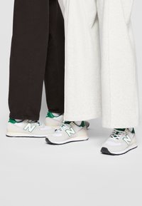 Tenisky New Balance s bílým a světle šedým designem s zelenými akcenty, texturovaným síťovaným svrškem, polstrovaným límcem a gumovými podrážkami.