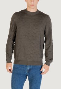 Dunkelbrauner Strickpullover mit einem strukturierten Wellenmuster, Rundhalsausschnitt und gerippten Bündchen. Kombiniert mit blauen Jeans.