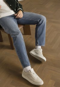 Baskets en daim gris clair avec des semelles blanches, portées avec un jean droit bleu. Personne assise sur une chaise en bois, sur un sol à motif chevrons.