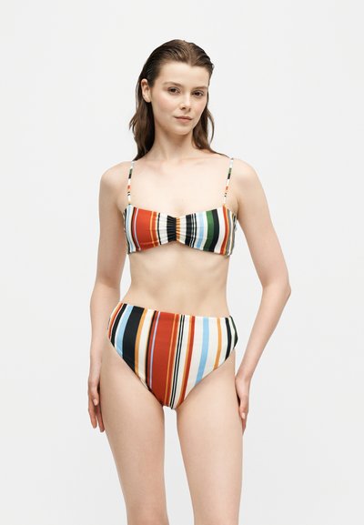 Jeune femme portant un bikini à rayures verticales multicolores avec un haut bandeau et un bas taille haute, debout devant un fond blanc uni.