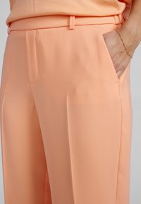 Béžové tailored nohavice s hladkým povrchom, ktoré majú bočné vrecká a rovný predný dizajn, doplnené o pútka na opasok.