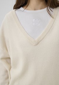 Pull en tricot couleur crème avec un col en V, superposé à un haut blanc côtelé avec un motif floral brodé sur la poitrine.
