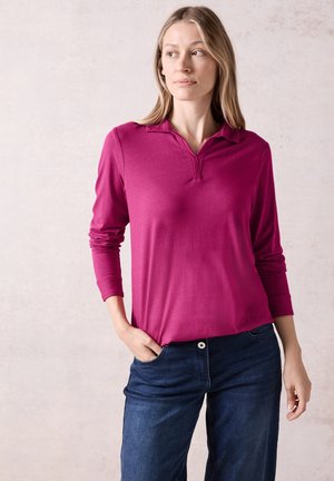 Langärmliges Poloshirt in leuchtendem Magenta mit einem kragenförmigen Ausschnitt. Kombiniert mit blauen Denim-Jeans, die über vordere Taschen und einen Knopf verfügen.