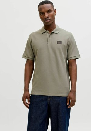 Vīrietis valkā īsroku olīvu zaļas krāsas polo kreklu ar mazu krūšu logotipu un tumši zilas džinsas, stāvot pret vienkrāsainu gaišu fonu.