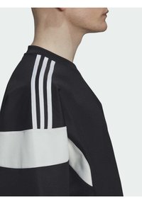 Mustasporttihuppari valkoisilla yksityiskohdilla, jossa on kolme valkoista raitaa olkapäällä ja teksturoitu kangas. Pyöreä kaula-aukko ja löysä istuvuus.