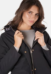 Giacca nera impermeabile con zip frontale, accessori in oro e cappuccio. L'interno ha una texture liscia e una fodera grigia.