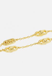 Brazalete de metal en tono dorado con eslabones ovalados entrelazados, un charm de logotipo central y acentos redondeados para un diseño moderno y elegante.