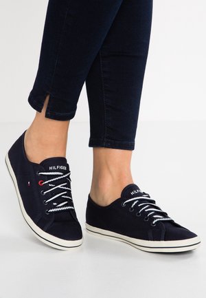 Osoba nosząca ciemnoniebieskie niskie trampki z białymi podeszwami i sznurowadłami, w połączeniu z ciemnoniebieskimi spodniami do kostek z rozcięciami przy kostkach.