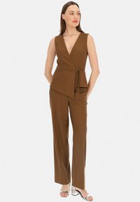 Brązowy bezrękawnik z głębokim dekoltem w kształcie litery V i bocznym wiązaniem, w zestawie z dopasowanymi brązowymi spodniami typu straight-leg i srebrnymi sandałami na obcasie.